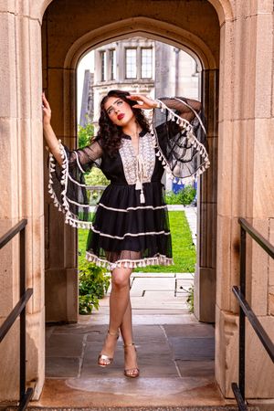 Woman in black boho dress with tassel cape poses elegantly under stone archway of historic building.
