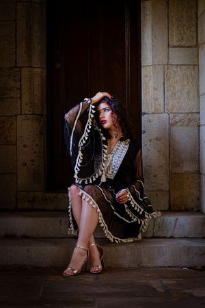 Woman in black embroidered boho dress with gold accents sits on stone steps by a wooden door.