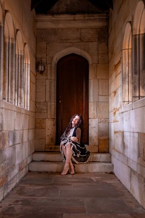 Woman in black dress poses on stone steps before arched wooden door in elegant gothic stone building.