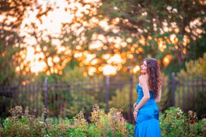 A woman in a blue dress stands in a sunlit garden at golden hour with bokeh trees in the background.