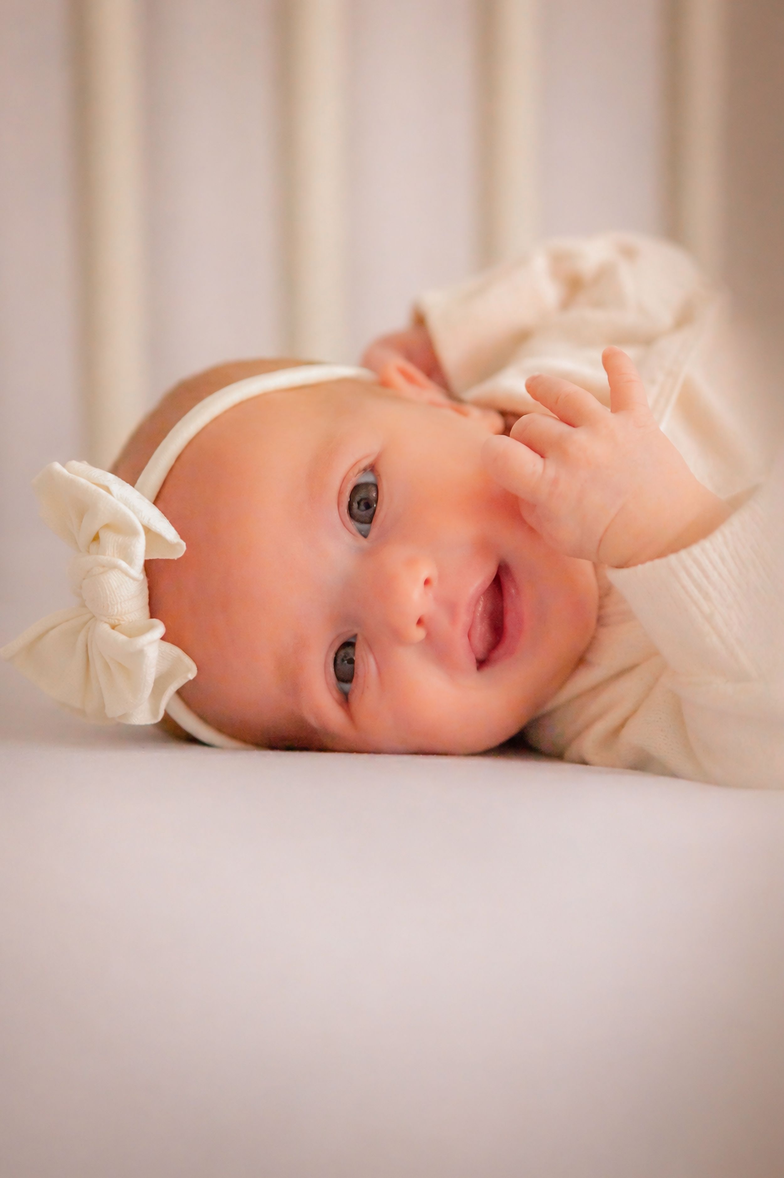 Smiling baby girl in cream outfit and white bow headband lying in a white crib, looking up with bright blue eyes.