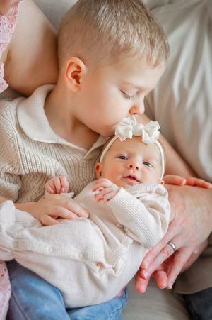 Young boy gently kissing newborn baby sister wearing white bow headband, both dressed in cream outfits.