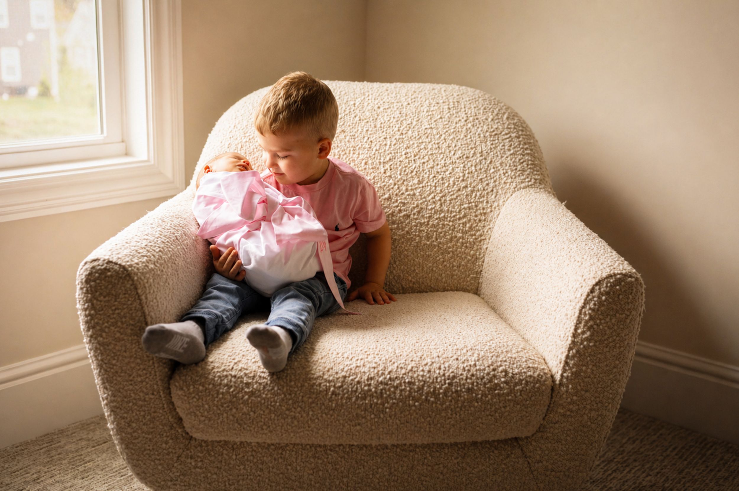 A young toddler boy sits in a cozy beige armchair holding a baby doll near a sunlit window.