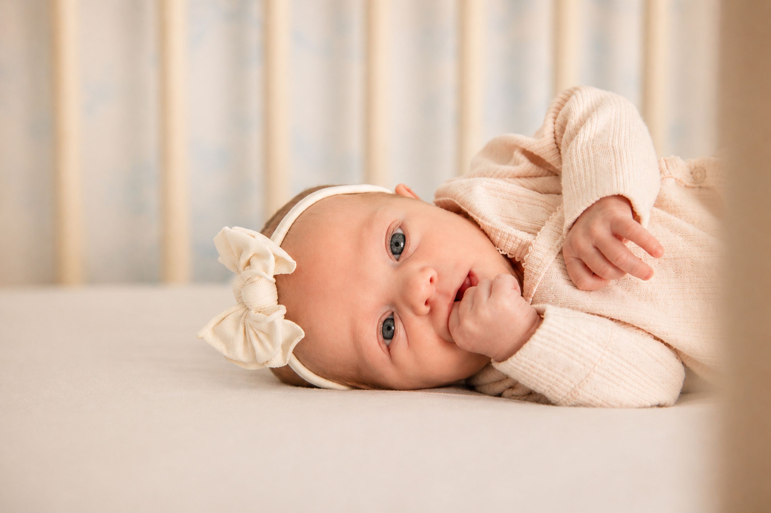 Newborn baby girl in pink outfit and white bow headband lying in a white crib with soft blue curtains.
