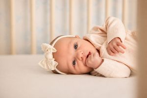 Newborn baby girl in pink outfit and white bow headband lying in a white crib with soft blue curtains.