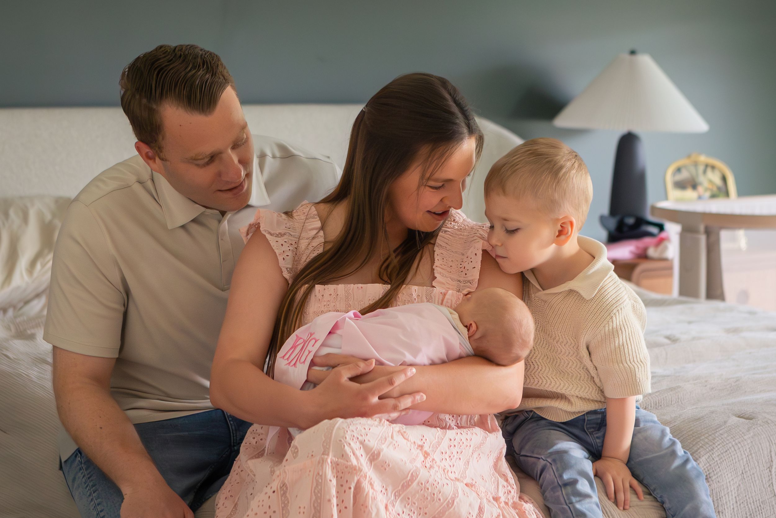Family of four with newborn baby girl in pink, parents and toddler son bonding on bed in soft natural light.