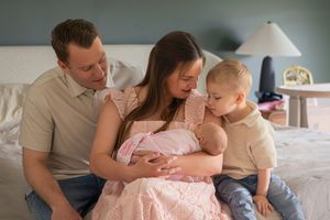 Family of four with newborn baby girl in pink, parents and toddler son bonding on bed in soft natural light.