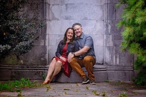 Happy couple sitting together against a stone wall surrounded by lush greenery in an outdoor engagement photo.