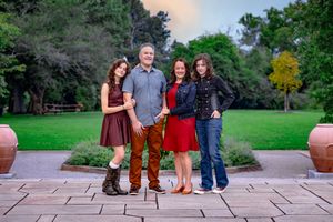 Family of four posing outdoors on a stone patio surrounded by lush green trees and garden landscaping.