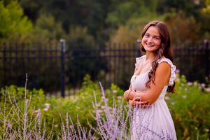Smiling young woman in white dress among lavender flowers in a garden with iron fence.