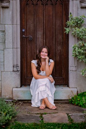 Young woman in white ruffled dress sitting on stone steps before ornate wooden door, smiling gracefully.