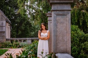 Young woman in white dress posing gracefully beside stone pillars in a lush garden setting.
