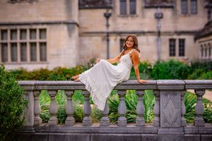 Young woman in flowing white dress sitting gracefully on stone balustrade outside historic building.