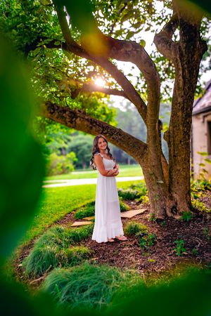 Woman in white dress standing under a large tree in a sunlit garden, surrounded by lush green foliage.