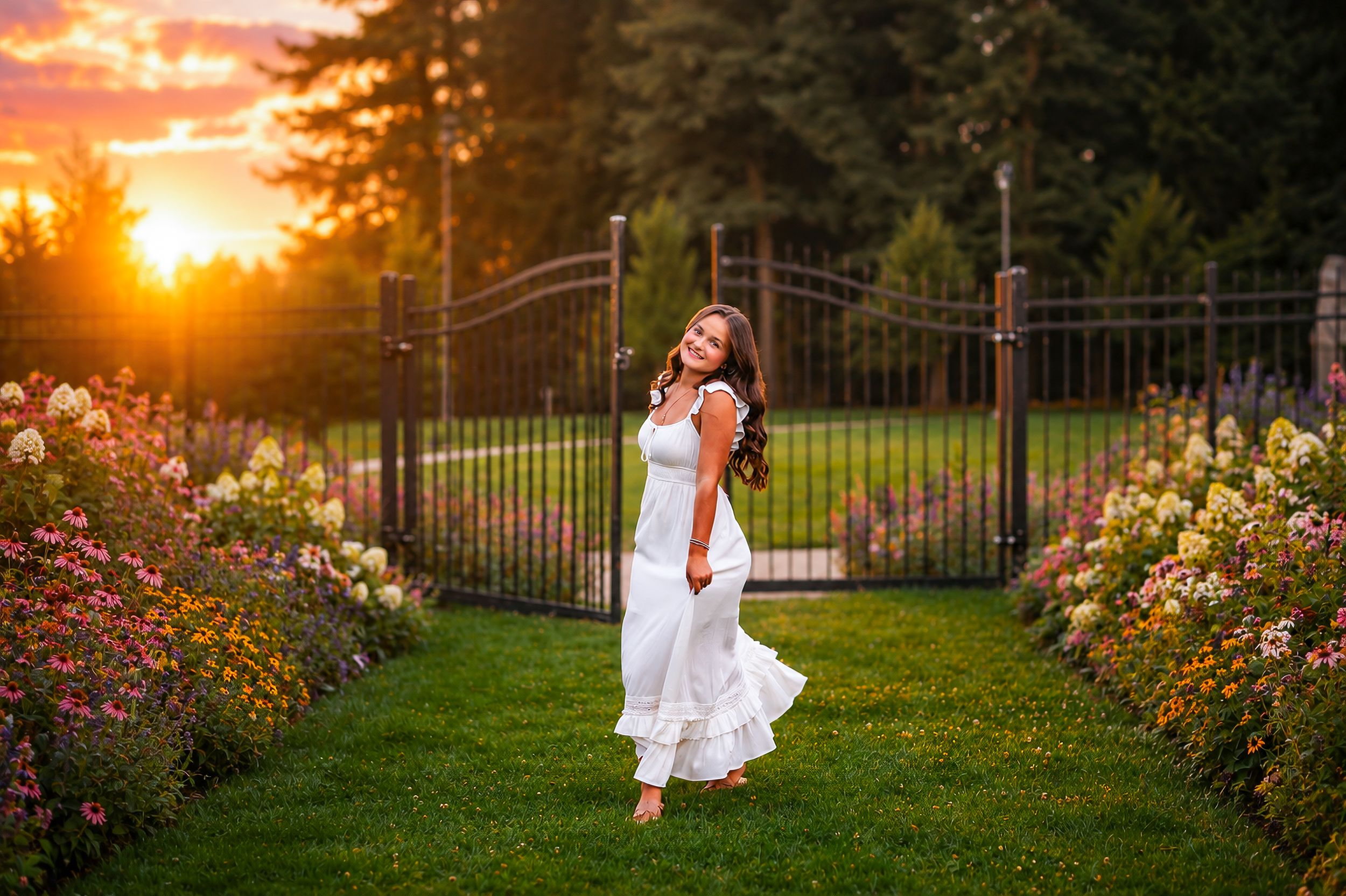 Woman in white dress standing in a flower garden at golden hour sunset with iron gate backdrop.
