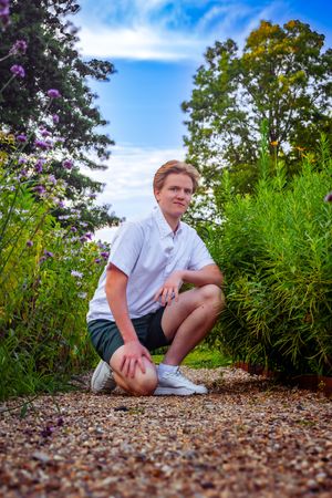 Young man in white shirt and green shorts crouching on a gravel path surrounded by lush garden greenery.