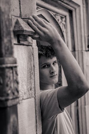 Young man leaning against ornate stone archway, black and white portrait photography.