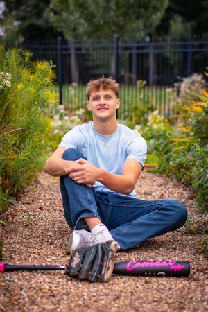 Young man in blue t-shirt and jeans sitting on gravel path with baseball glove and pink bat nearby.