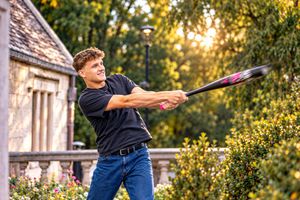 Young man in black shirt swinging a baseball bat outdoors with golden sunlight and green trees in background.