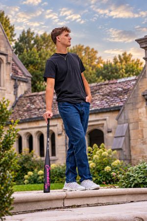 Young man in black t-shirt and jeans holding a baseball bat stands on stone steps before a historic building at dusk.