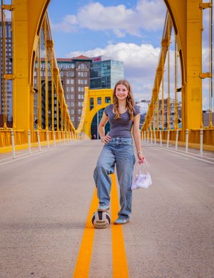 Young woman poses with soccer ball on yellow Pittsburgh bridge, wearing jeans and gray tank top, city skyline behind her.