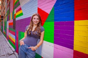 Young woman smiling in jeans and gray top, leaning against vibrant colorful mural wall in urban alley.