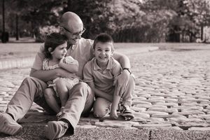 Black and white photo of a bald father sitting on cobblestones with two smiling children in a park.