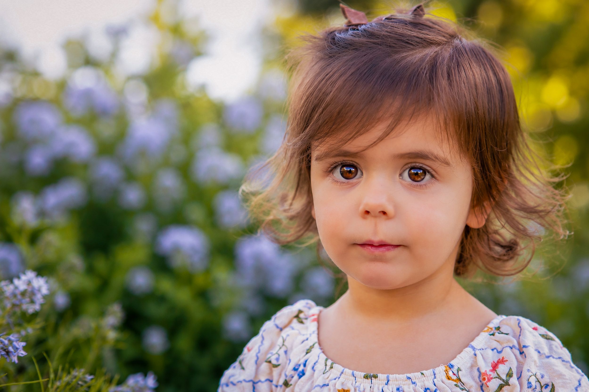 Adorable toddler girl with brown eyes and short hair posing outdoors near purple wildflowers in soft natural light.