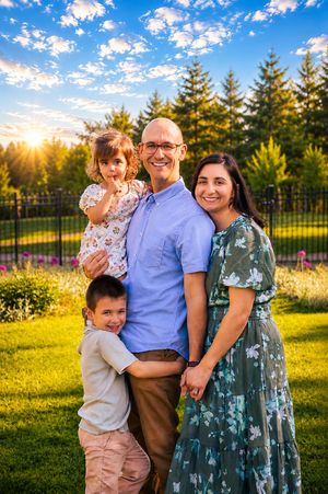 Happy family of four poses outdoors at golden hour with lush green trees and colorful flowers in the background.