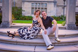 A smiling couple in elegant attire sits on stone steps outside a classic building with lush greenery.