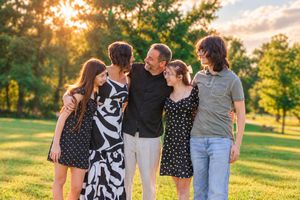 Family of five smiling and embracing outdoors at golden hour in a lush green park.