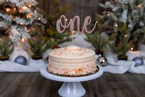 First birthday cake with rose gold 'one' topper on white cake stand, surrounded by snowy pine trees and ornaments.