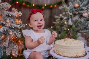 Happy baby girl in white dress with red bow smiles beside a frosted Christmas tree and white cake.