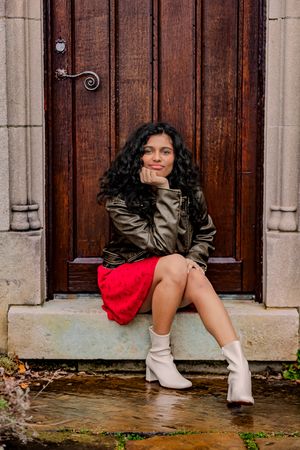 Young woman with curly hair in red skirt, leather jacket, and white boots sitting on stone steps by a wooden door.