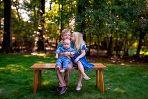 Three young siblings sitting on a wooden bench outdoors, older girl kissing brother's cheek while holding baby.
