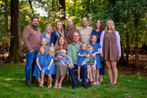 Large multigenerational family posing outdoors in coordinated blue and neutral outfits surrounded by lush green trees.