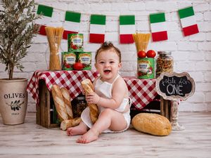 Baby smiling at Italian-themed first birthday cake smash with bread, tomatoes, pasta, and red checkered tablecloth.