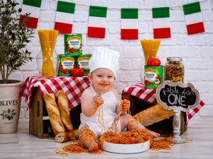 Baby in chef hat sits in spaghetti for first birthday smash cake photo with Italian-themed decorations.