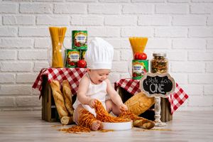 Adorable baby in chef hat plays with spaghetti surrounded by Italian ingredients and bread for first birthday.