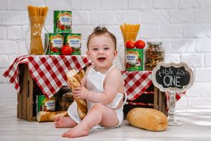 Smiling baby in white onesie sits surrounded by Italian food props, pasta, bread, and canned tomatoes with a Table for One sign.