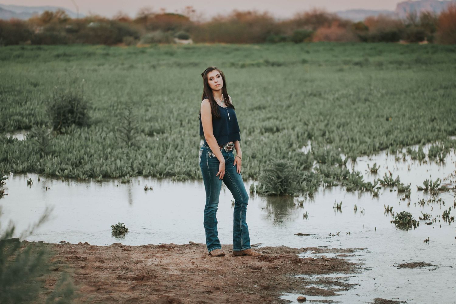 Person in dark top and jeans standing by reflective water in green field at sunset.
