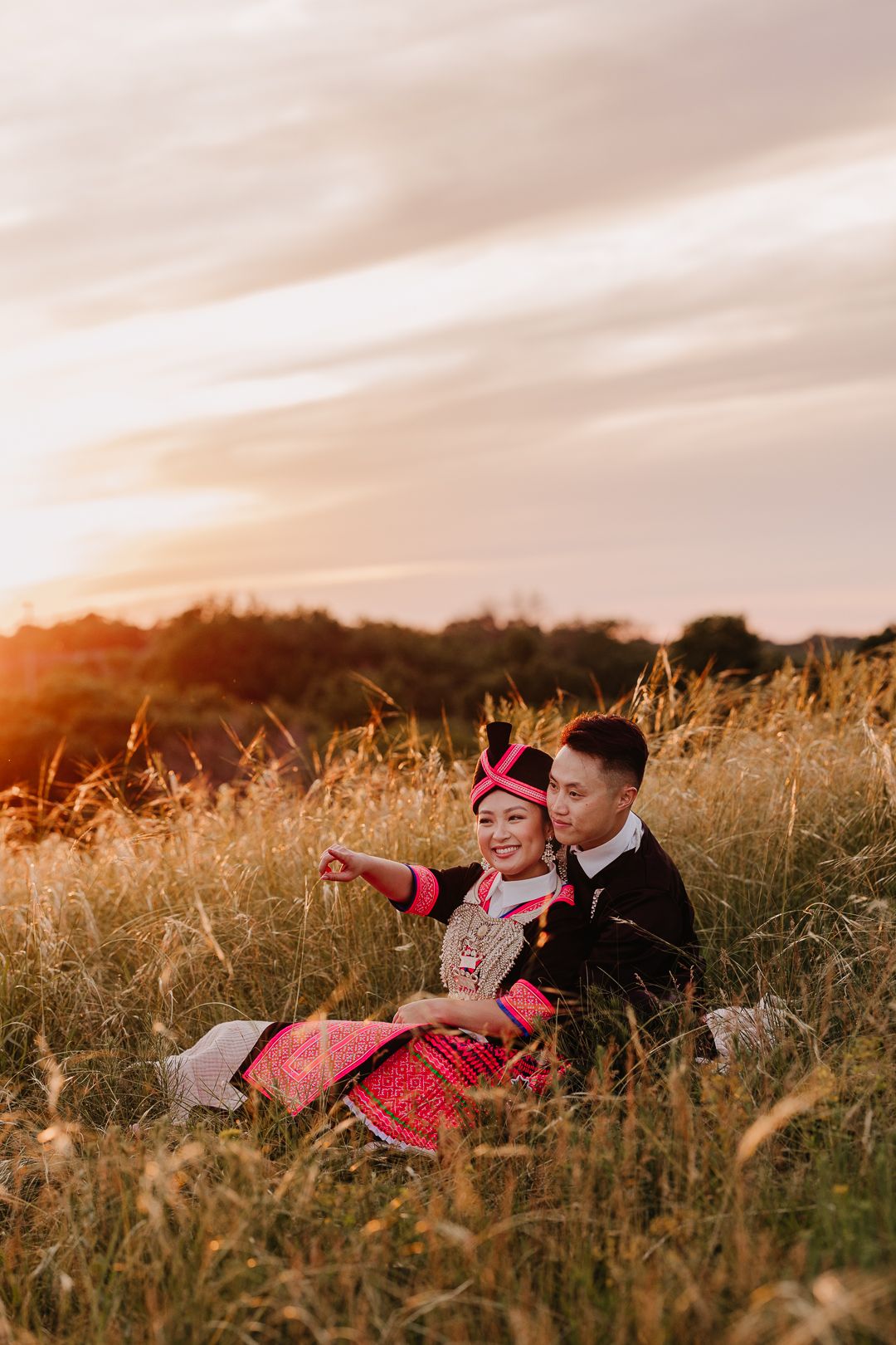 A couple in traditional Hmong attire sits in a golden field during sunset.