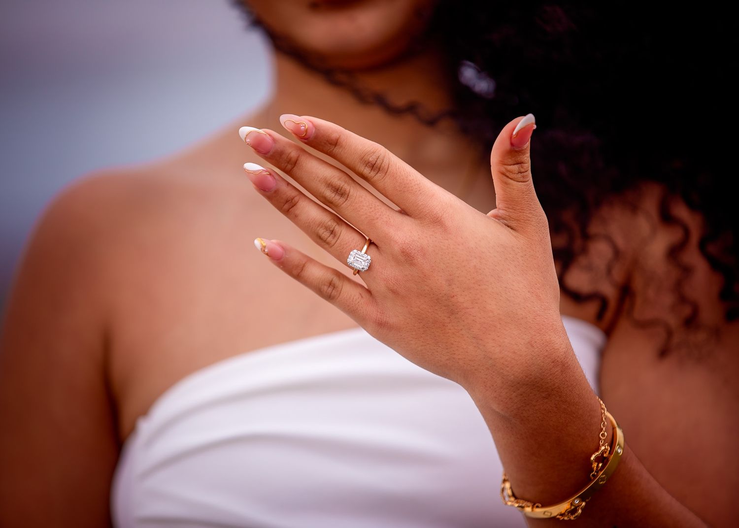 Hand wearing elegant diamond engagement ring and gold bracelet against white fabric background.