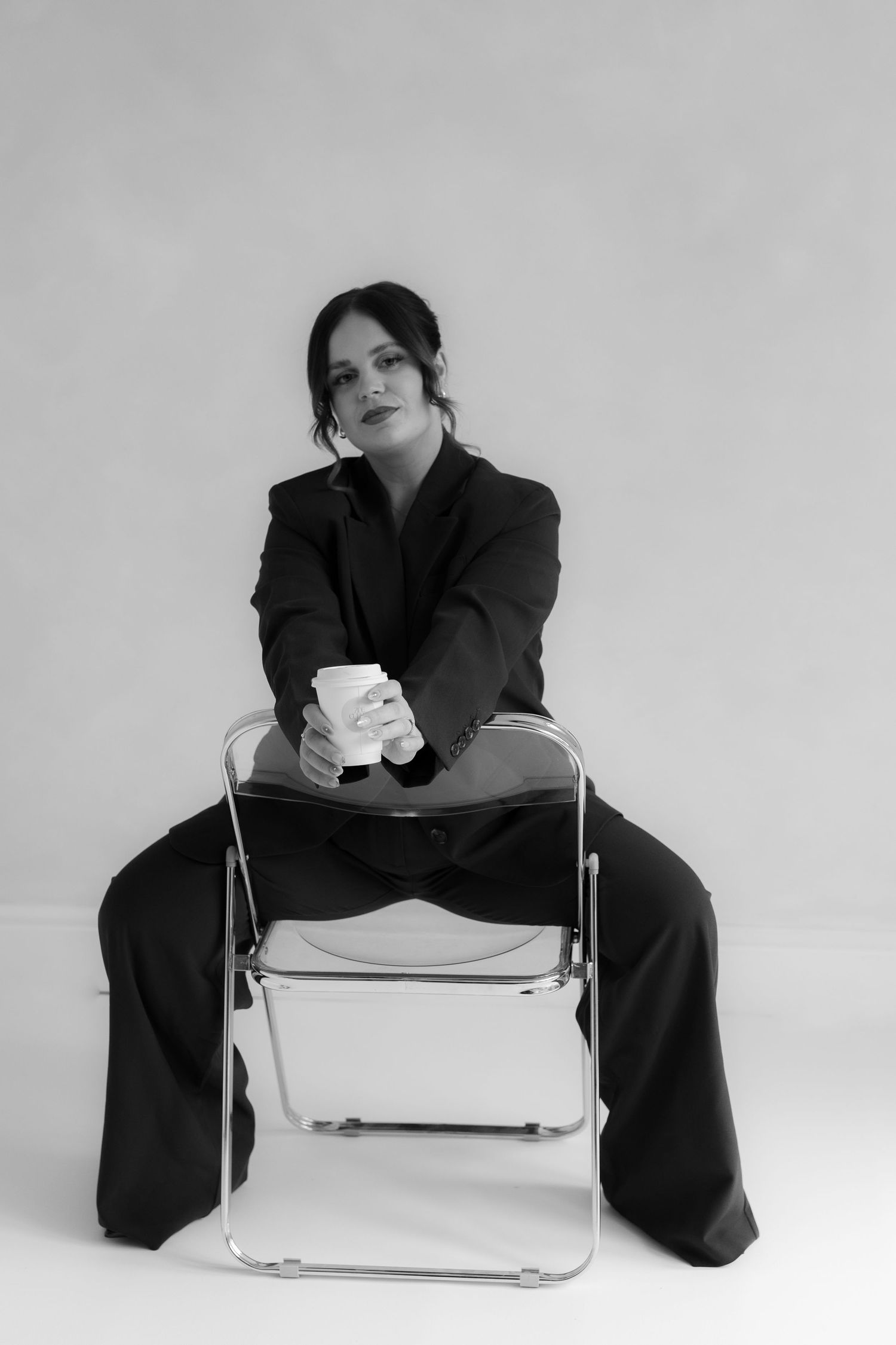 black and white image of photographer branding session while sitting backwards on a clear chair holding a coffee in studio setting.