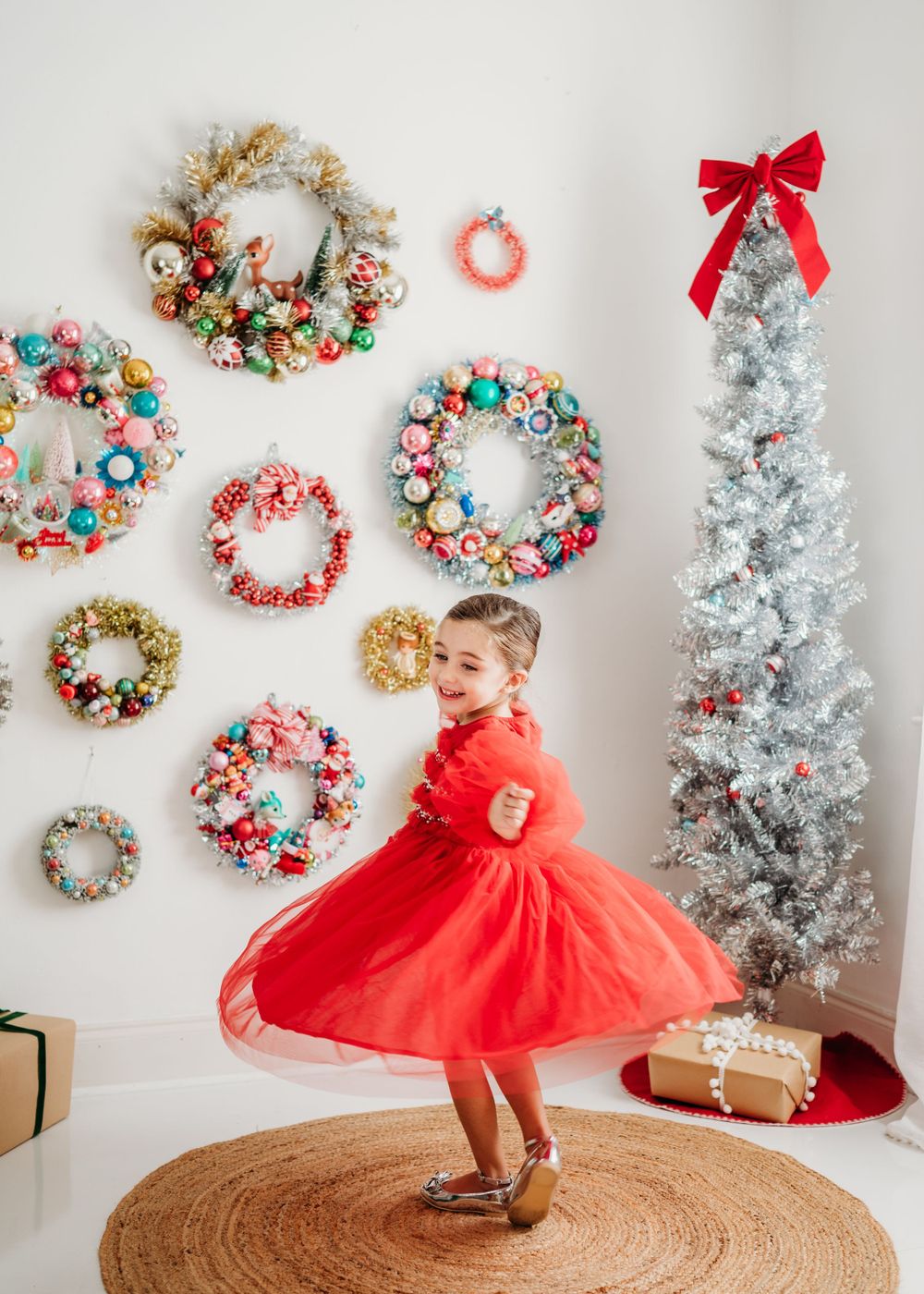 Child twirls in red holiday dress against white wall adorned with colorful vintage ornament wreaths and silver Christmas tree.