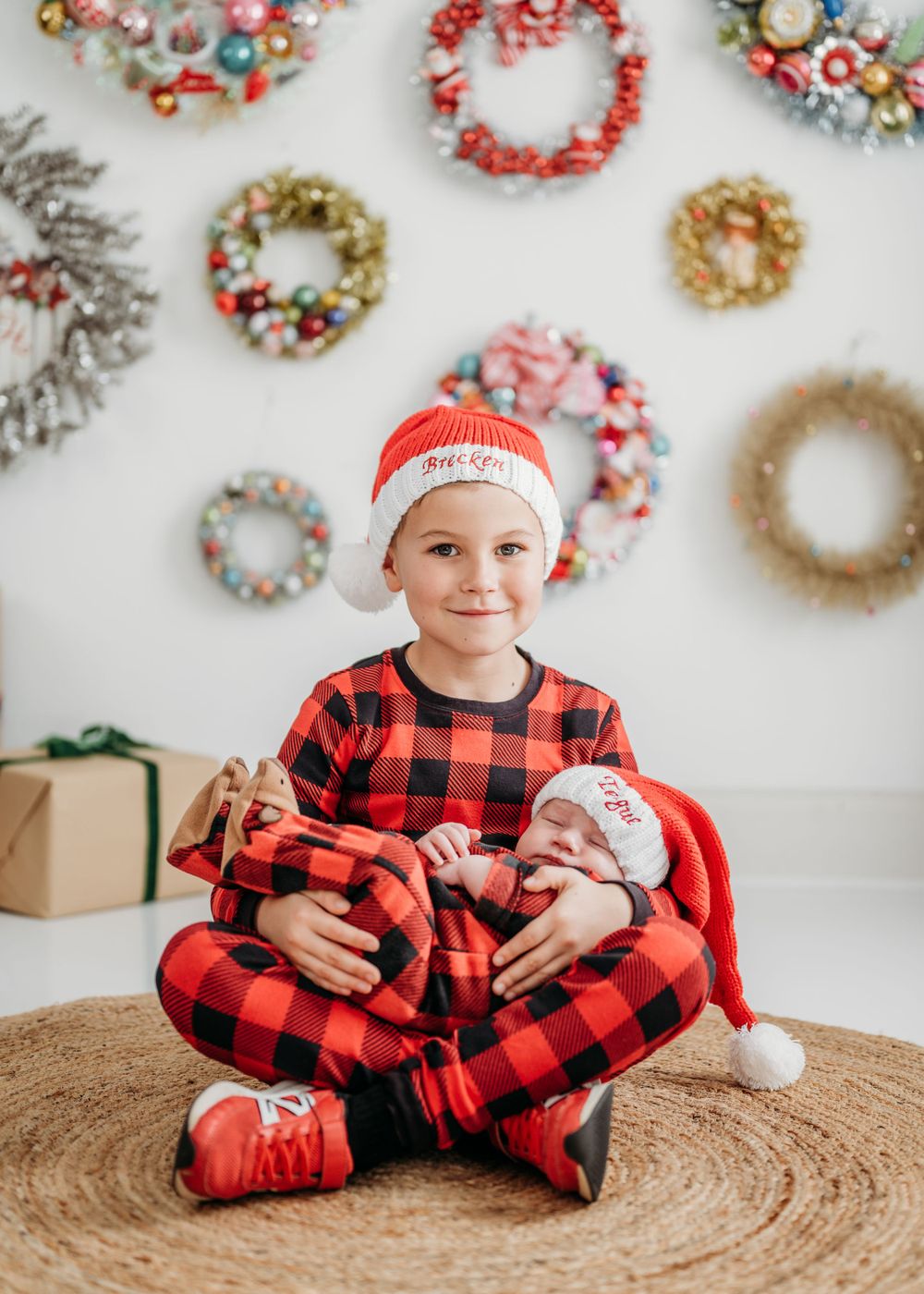 A buffalo plaid Christmas scene with decorative wreaths on white wall and woven rug on floor where a big brother holds his new baby sister.