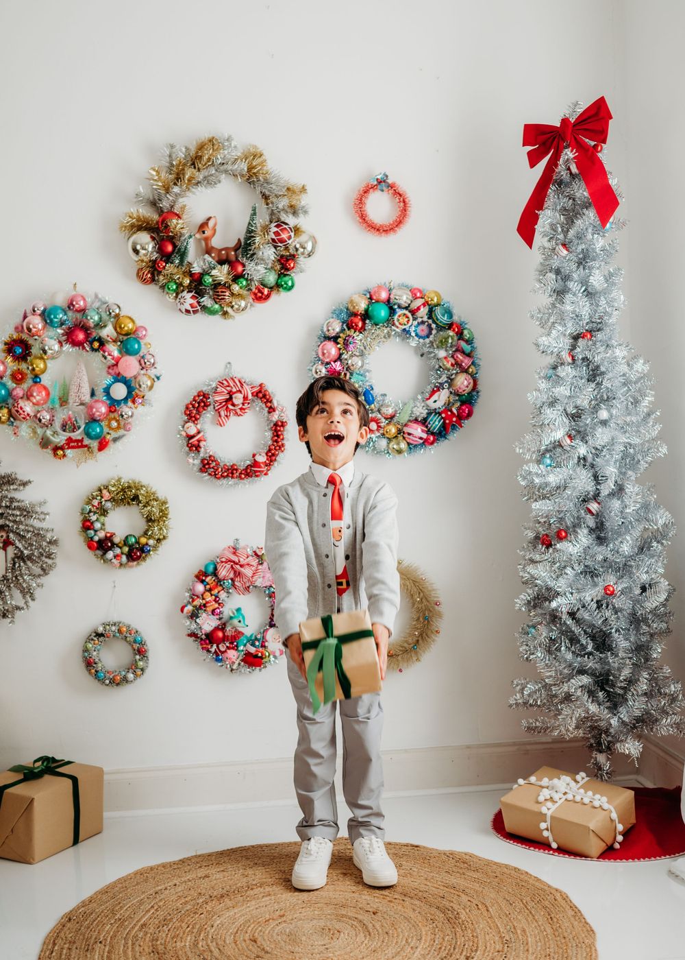 A stylish holiday scene with decorative vintage christmas wreaths on white wall, flocked Christmas tree, and wrapped presents on jute rug.