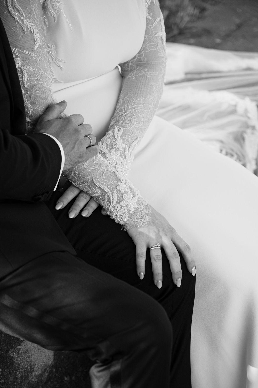 Close-up black and white detail of delicate lace sleeves and hands resting on a white wedding dress.