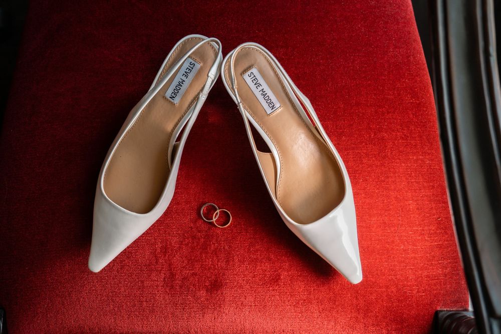 White bridal heels and wedding rings displayed on red velvet background in elegant arrangement.