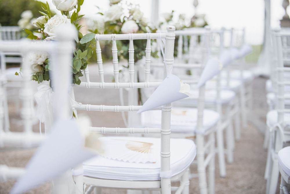 White wedding ceremony setup with chairs and floral arrangements in an elegant outdoor garden setting.